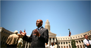 A photograph of Mayor Hancock, an African American man, in his 30s-40s, wearing a suit. He is standing outside of a government building.