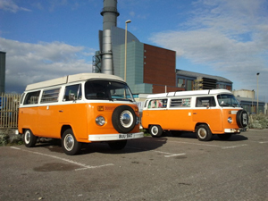 A photo of two vintage, orange and white Westphalia VW vans parked side by side
