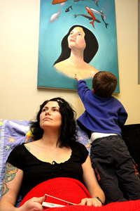 Photo of a woman sitting on a sofa looking off to the left. Above here hangs a painting of another woman who has similar feature and is also looking off to the left. 