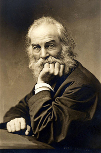 A sepia tone photo of Whitman looking at the camera with one hand under his chin and the other resting on a table. He has a long beard and mustache and big, light-colored eyes.