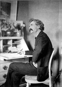 Mark Twain seated at his desk, looking upward as if in thought. He seems to have been interrupted while writing. The room shown in the old black and white photograph seems bare, but there are several vases filled with flowers on Twain's desk.