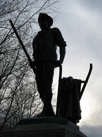 A photograph of the Minute Man monument in Concord, Massachusetts. It is a man holding onto a plow with one hand and a musket in the other.