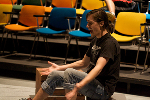 photo of a seated woman in casual clothes rehearsing a scene from a play in an empty theater; her arms stretch outward, palms are face up, and her face expresses a serious mood