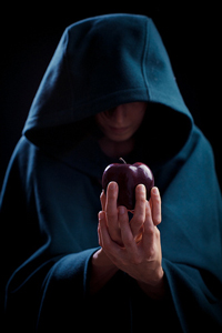 photo of someone portraying a witch. This person wears a dark cape; only part of their face and their hands show; and an apple is held out to the viewer.