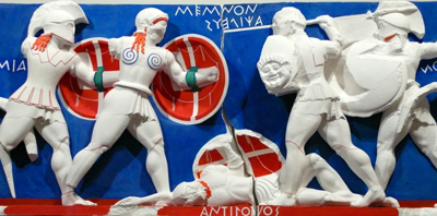 A plaster relief showing classical Greek warriors fighting. Some have shields and helmets. There are labels in ancient Greek written into the background.