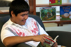 A photograph of a male student sitting in a chair in a library, reading a book