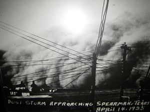 black and white photo of mountain-high clouds of dust just before rolling over several farm buildings; telephone and electrical wires stretch in different directions from leaning poles in the foreground; handwriting on photo reads ‘Dust storm approaching Spearman, Texas April 14, 1935’