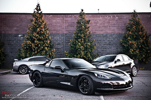 Photo of three Corvettes in a parking lot, two black and one silver. Can only see the one in the foreground completely. Backdrop is a brick wall with three Christmas-shaped fir trees