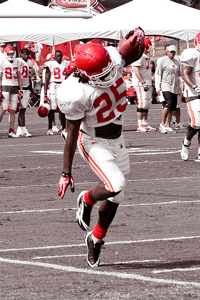 A football player at practice. The image is black and white except for the red of the player's uniforms. The camera has caught a player balancing on one foot, almost like he's hanging in the air.