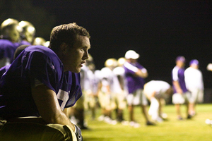 A player resting after a scrimmage in a football game. The image is somber and dark, except for the player's purple jersey.