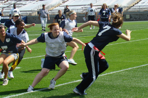 photo of young women playing flag football on a football field. Coaches and an umpire looks on in the background.