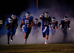 A football team charges out of a cloud of blue and white smoke