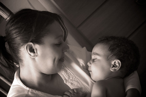 A photograph of a mother holding a sleeping baby. She is looking down lovingly at the baby.