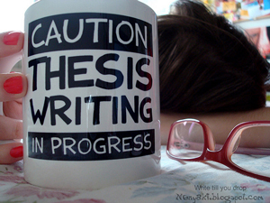 photo of a writer with head on a desk and coffee mug and reading glasses in the foreground. The mug reads “Caution Thesis Writing in Progress.”