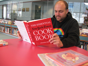 A photograph of a man reading “The Essential New York Times Cook Book” in a library