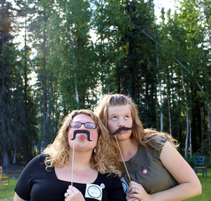 A photograph of two young women posing for a photo with fake moustaches.