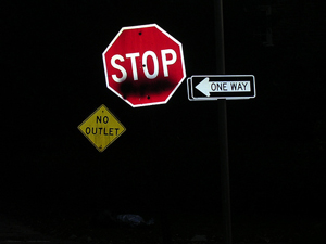 A photograph of confusing signs. One reads “Stop”, the other “One Way”, and the other “No Outlet.”