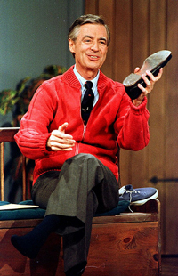 A photograph of Fred Rogers on the set of his show “Mr. Roger's Neighborhood.” He is sitting on a bench and changing his shoes.