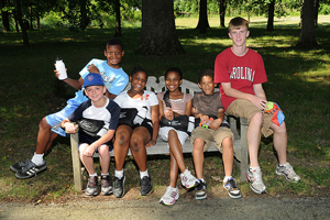 A photograph of a group of boys and girls with their camp counselor sitting on a bench and smiling.