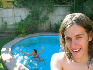 A photograph of two teens. One, the female is in the pool, the male is outside the pool close to the camera.