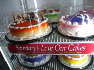 A photograph of cakes in a cooler. The is a sign between them that reads; “Secretary's love our cakes.” There is an apostrophe in Secretary's which is an incorrect use of this punctuation.