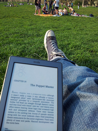 A photograph of a person reading a Kindle reader in a park. One of their feet is visible