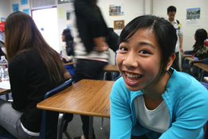 A photograph of a girl in a classroom sitting at her desk smiling