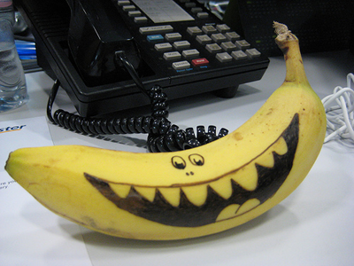 An image of a desk with a telephone, papers, and other office supplies. At the front of the photograph is a bright yellow banana with a huge, smiling face drawn on its side.