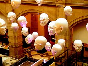 A large display of art in a Scottish museum. The museum itself is very formal, with dark red and gold hardwood and an elegant patterned tile floor. High up in the air, a mobile of dozens of white plaster floating heads float through the air on strings, each one showing a different exaggerated facial expression and a different emotion. 
