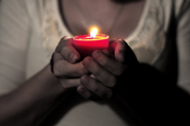 A black and white photograph of a woman cradling a lit candle in her hands. The candle is shown in color, and radiates a warm light.