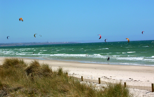 A beach with blue skies and deep blue-green water. Many hoop-shaped kites fly in the wind.