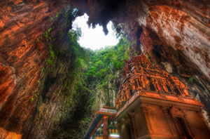 A photograph from below of a decorated, painted temple, a tall building covered in statues and decorations. It seems dwarfed by the what might be the mouth of a cave or extinct volcano overhead, a tunnel leading into sunlight, draped with vines and trees.