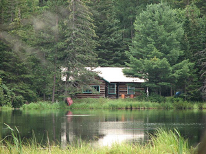 A photograph of a small cabin in the woods near the edge of a pond
