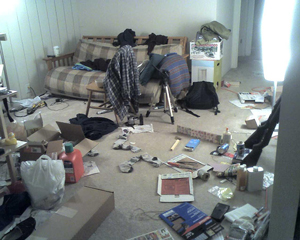 Photo of messy room: clothes strewn, paper and projects all across floor, stacked boxes, and other stuff