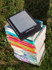 A stack of regular paper books, with an e-reader on the top. The stack of books is outdoors on the grass.