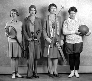 Four college aged women dressed in 1920's gym clothing and holding various sporting equipment.