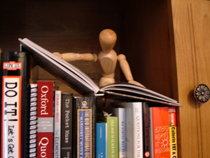 A close-up image of a bookshelf. A wooden artist’s mannequin stands behind the books, holding open one book as if reading it.