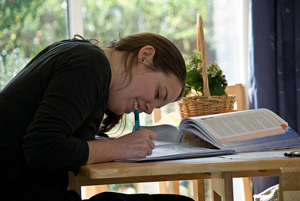 A photograph of a female student at a desk writing. She has a textbook open on her desk as well