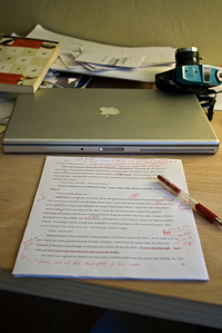 A photograph of a printed paper on a desk with a laptop, books, papers, and a camera. There is a pen laying on the printed paper, which has been marked by the pen.