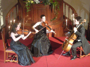 A photograph of a string trio consisting of two violins and a cello. The musicians, two women and a man, area all wearing formal attire.