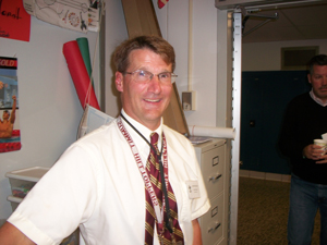 A photograph of a male teacher in a school. He is wearing glasses and has several lanyards hanging around his neck.