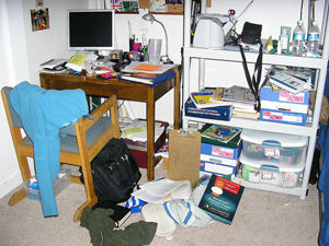 A photograph of a messy study area in a bedroom. In the area is a desk with a chair, a book bag, a computer, a desk lamp and a bookshelf. Strewn about the area are clothes, papers, notebooks, and other personal items.