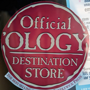 A sign in a store window that reads “Official 'Ology; Destination Store”