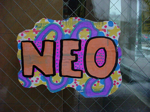 A photograph of the prefix “NEO” painted on cardboard which is hung on a fence