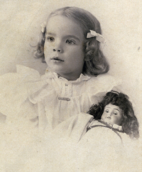 An older photograph of a young girl with a doll; They both appear to be wearing the same formal dress.