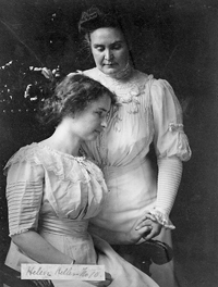 A photograph of Helen Keller holding hand of her teacher, Mrs. John A. Macy; Both are wearing formal looking dresses typical of the early 20th century.