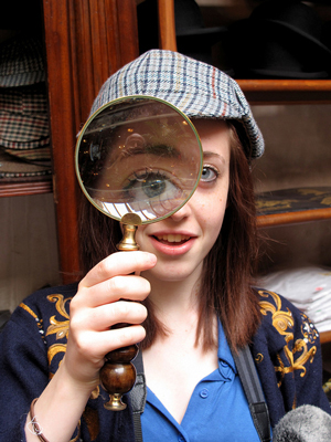 A photograph of a young woman looking through a magnifying glass. She is also wearing a ‘Sherlock Holmes’ hat