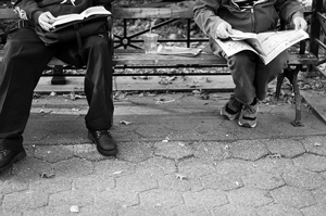 A photograph of two readers sitting on a park bench. The older reader is reading a book, while the younger reader is reading a newspaper.