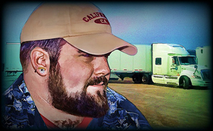 A photograph of a truck driver with his rig in the background. He is wearing a baseball hat.