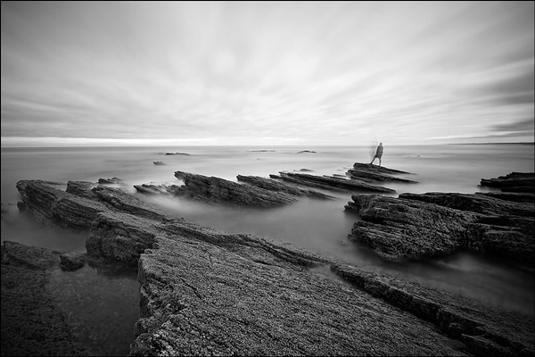 A monochrome photograph of a person standing on rocks at low tide